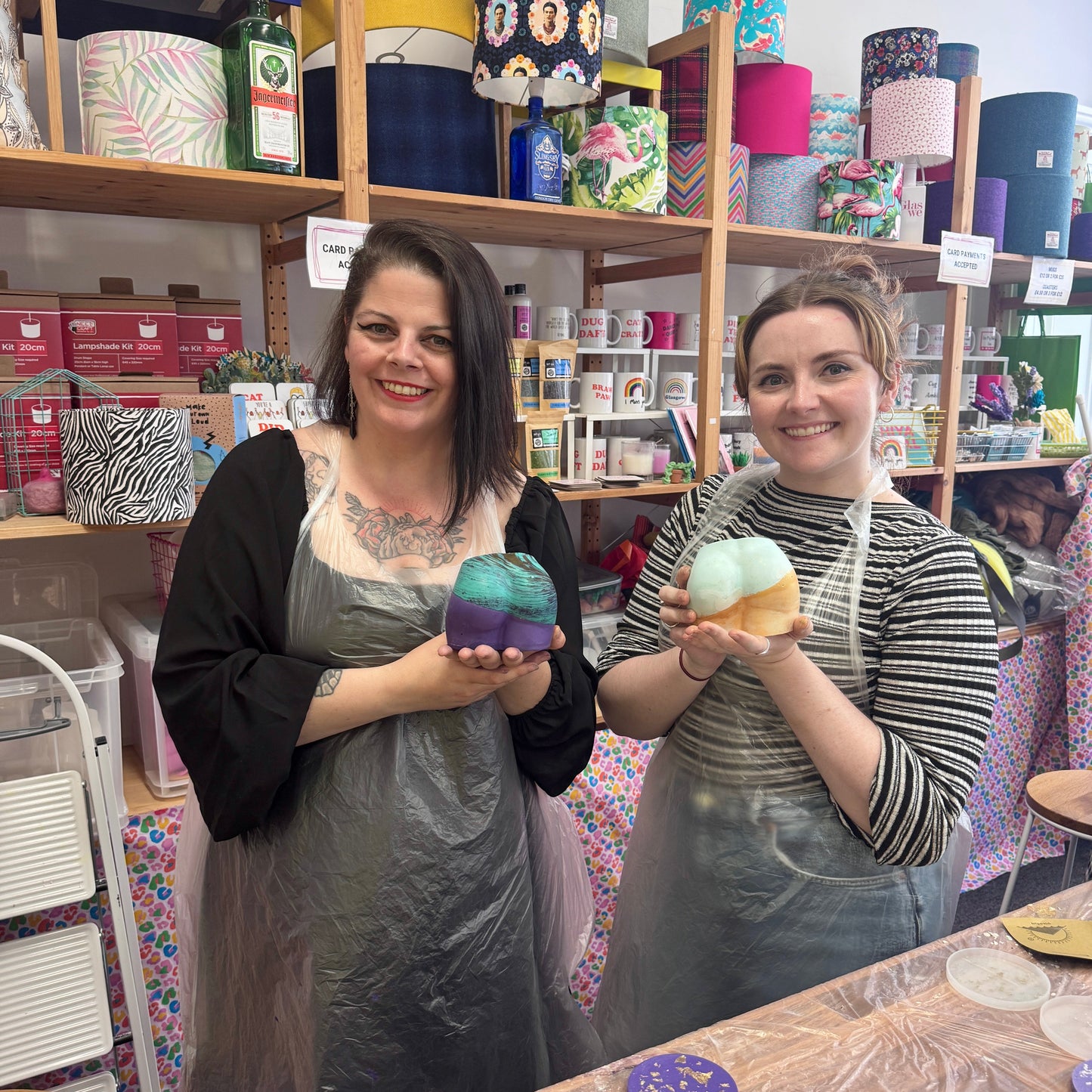 Two women holding colorful Jesmonite Bum Pots in a craft studio with shelves of finished products.