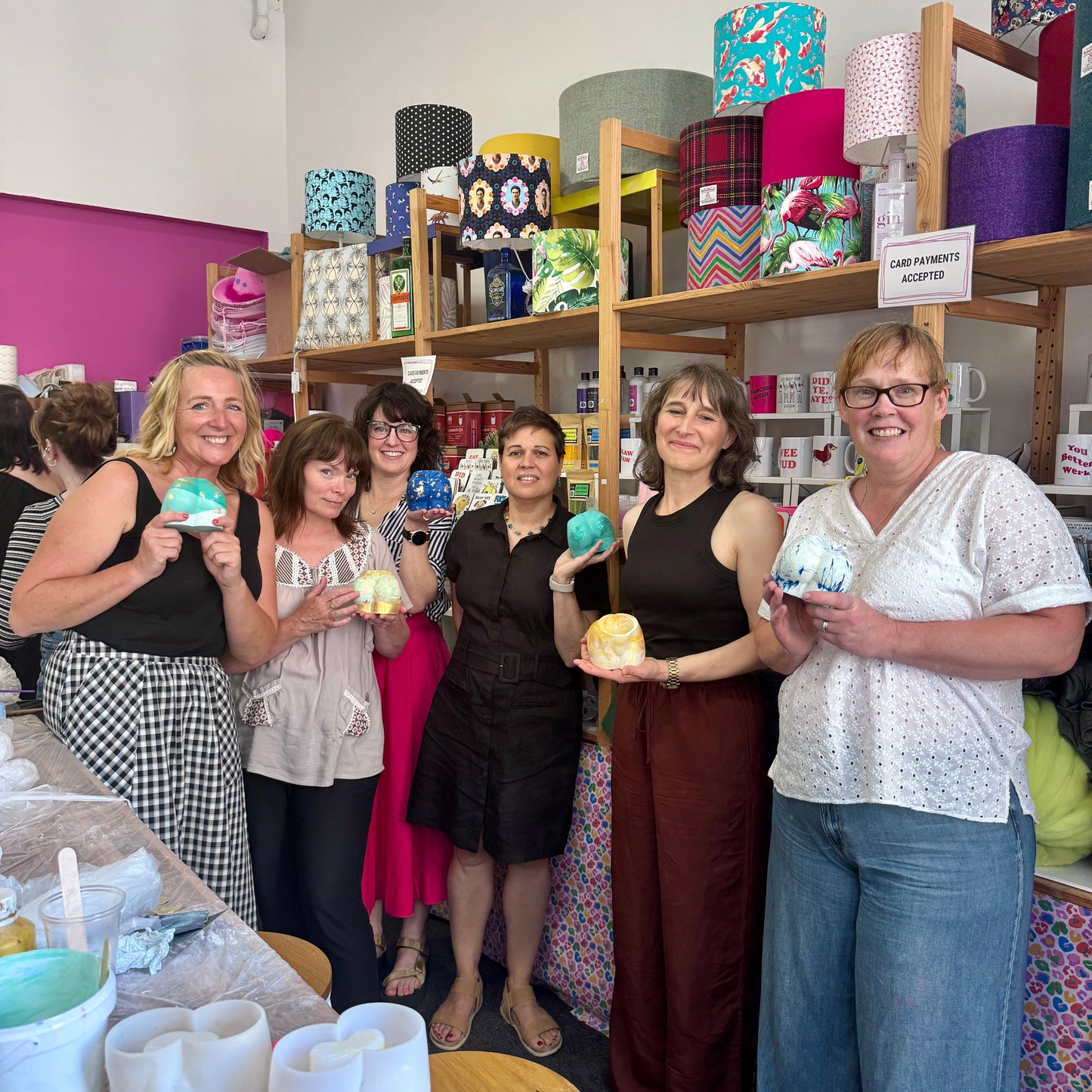 Group of women holding colourful Jesmonite Bum Pots in a craft studio with shelves displaying various items.